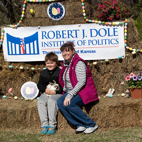 A boy sits on a bale of hay holding a rabbit in his lap while his mother sits next to him