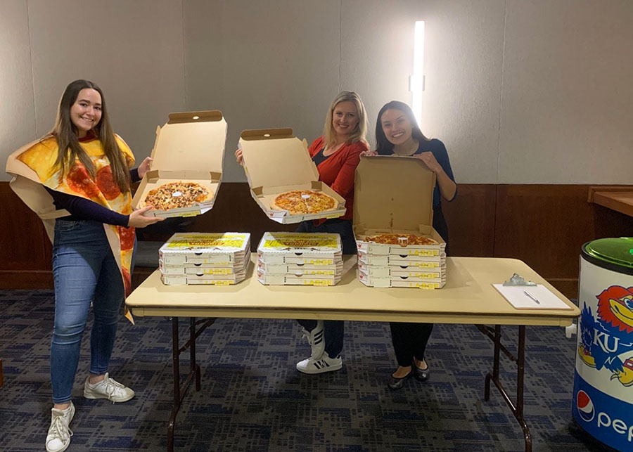 Three women pose with open pizza boxes next to a table stacked with pizza boxes.