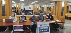People wave while sitting at a table with laptops, with signs reading "National Voter Education Week," and "Register to Vote today!"