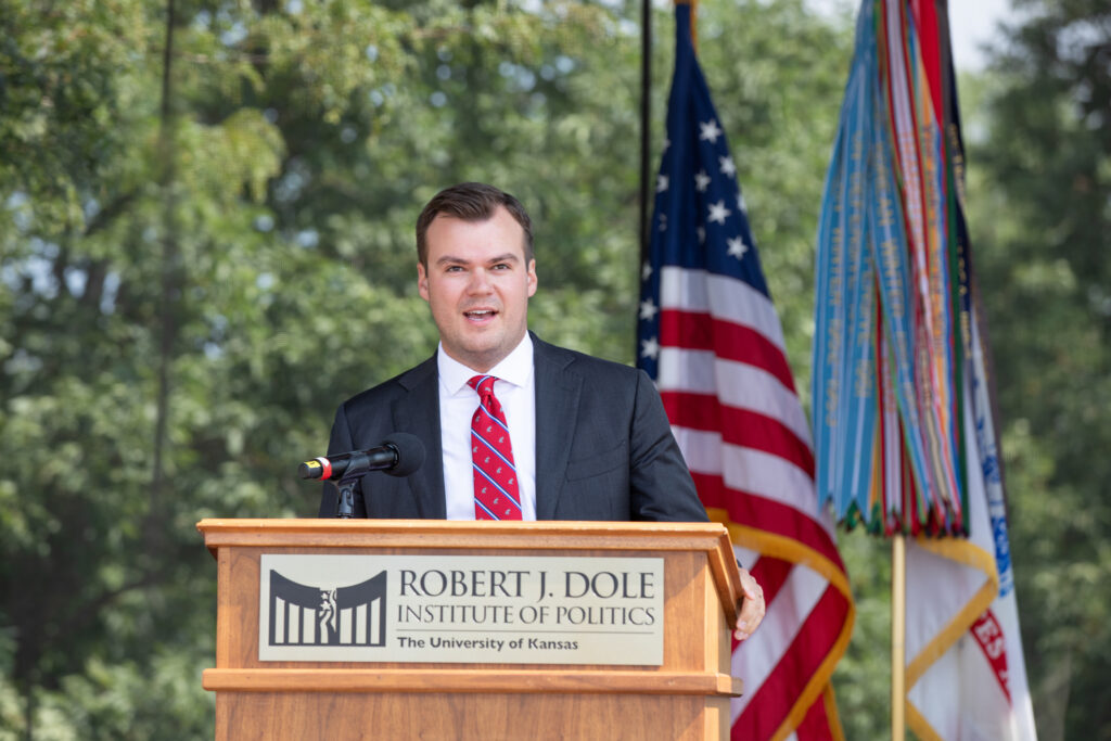 A man speaks from a podium on a stage with flags in the background.