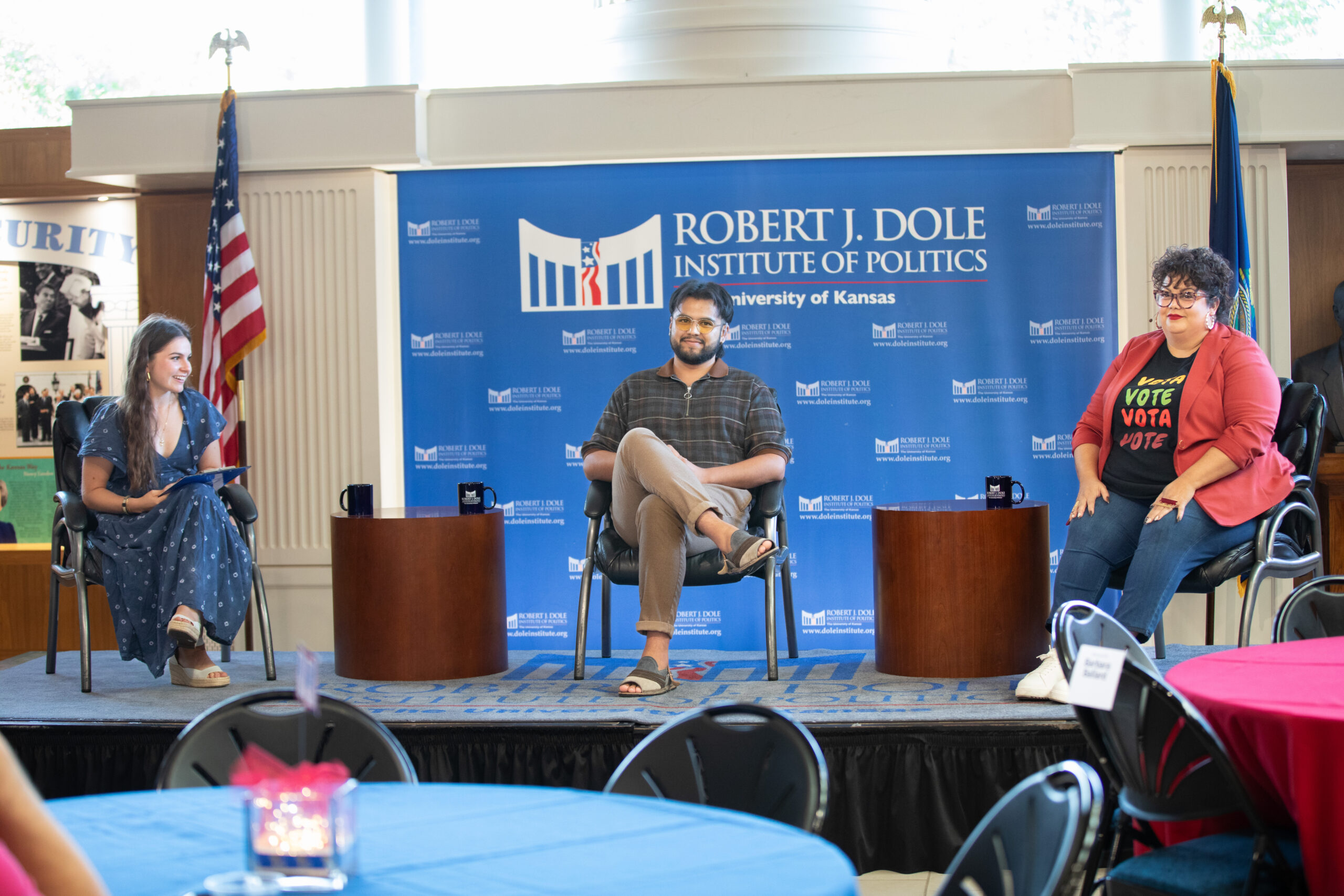 Three people look on as they sit on a stage with tables and chairs in the foreground.