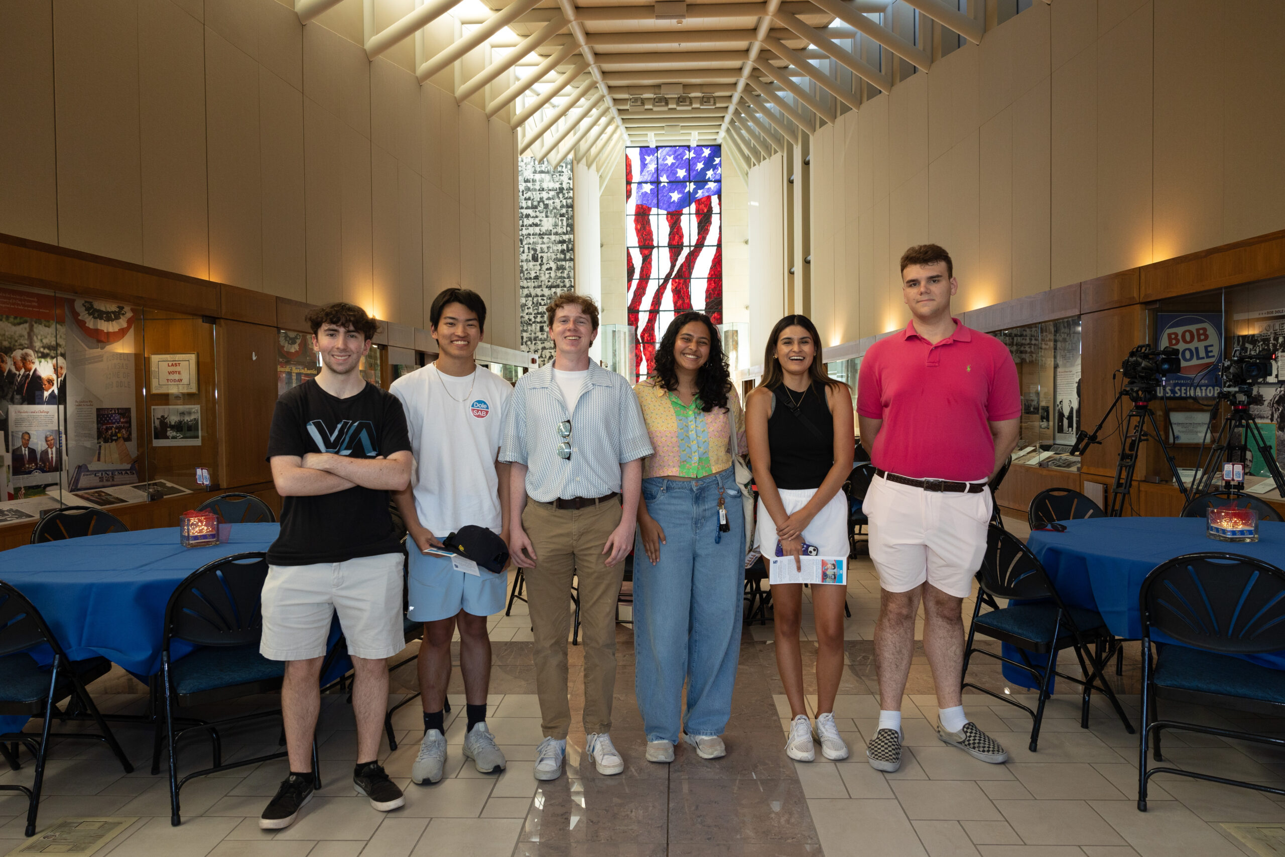 A group of men and women stand in the Dole Institute's main hall with a stained glass American flag window in the background.