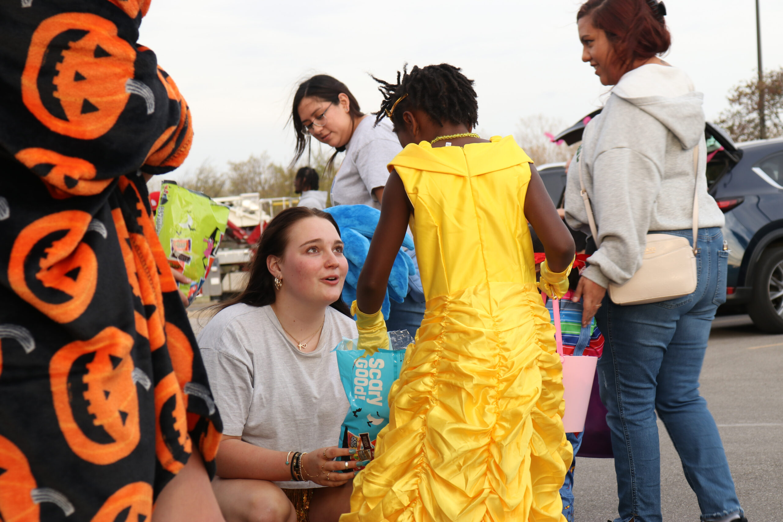 A woman kneels to give candy to a young woman in a Belle costume while others look on.