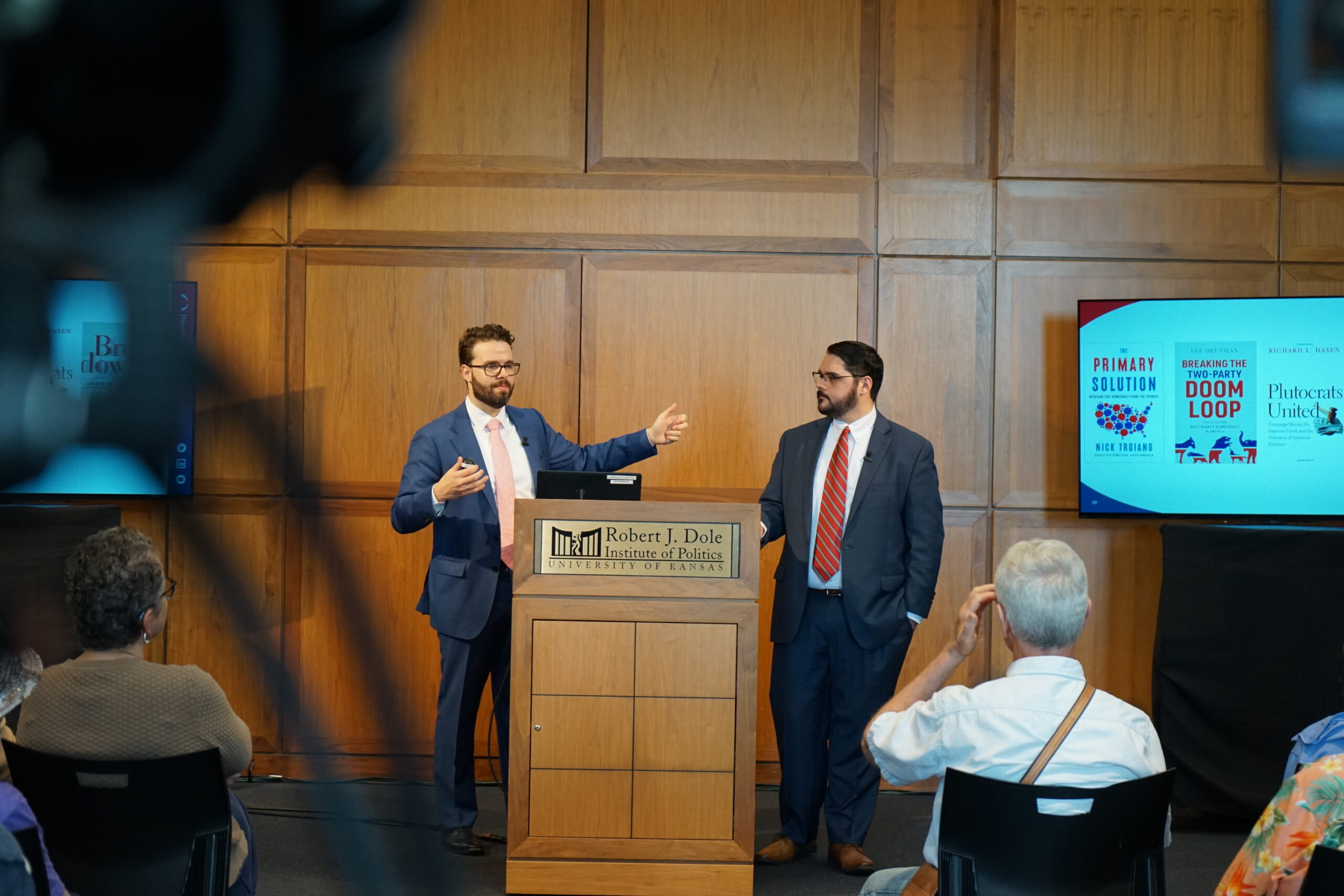 Two men stand at a podium in a large room with wood panels. One is gesturing while the other and members of the audience look on.