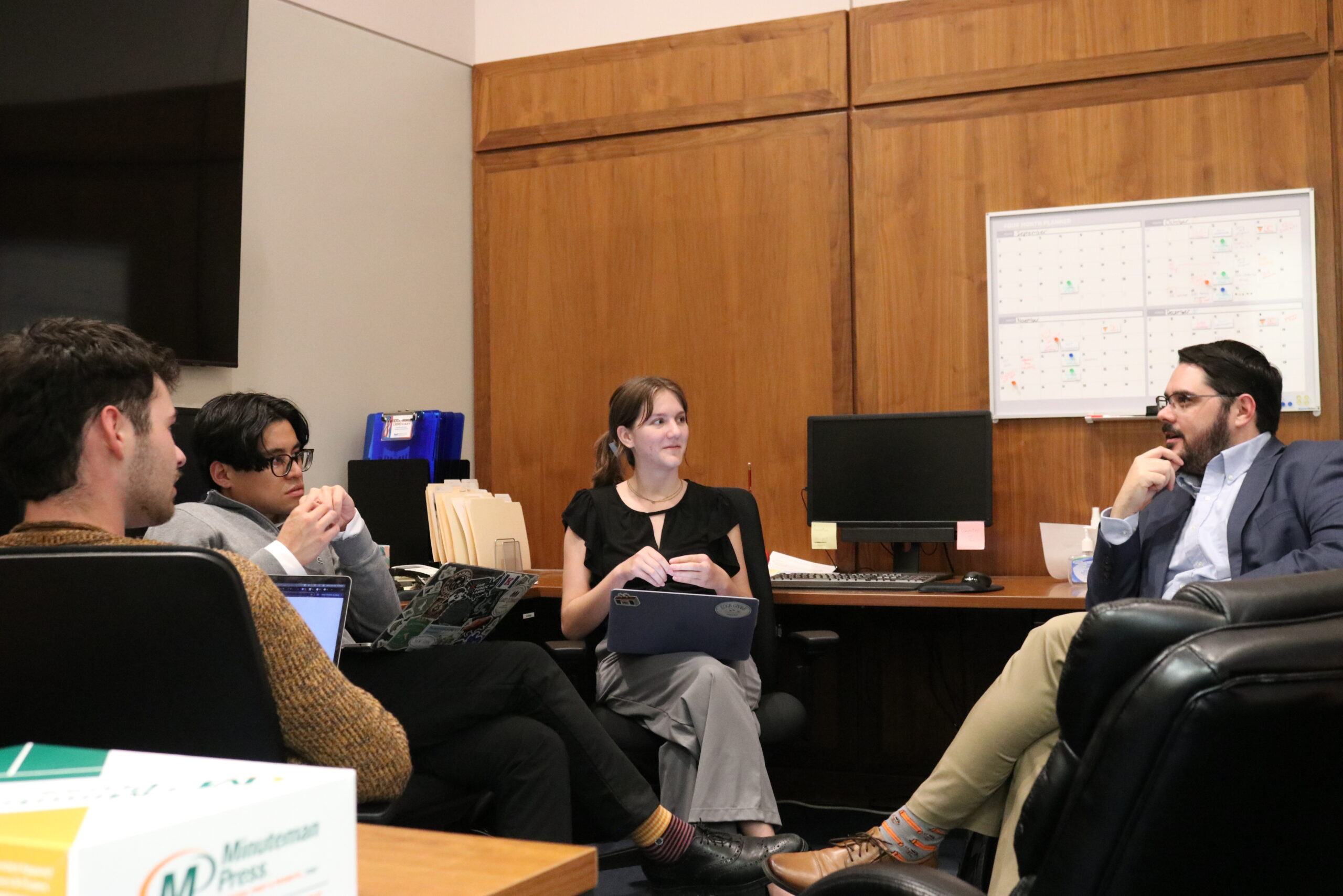 A group of people sit in a circle in an office area, with some holding laptops in their hap while listening to the conversation.