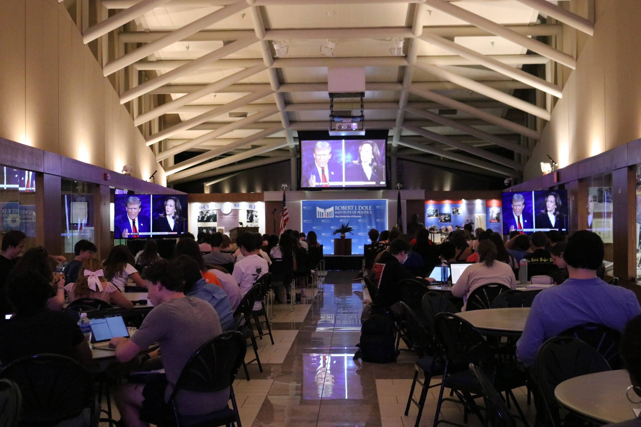 Students sit at tables in a large hall while watching a presidential debate on large screens.