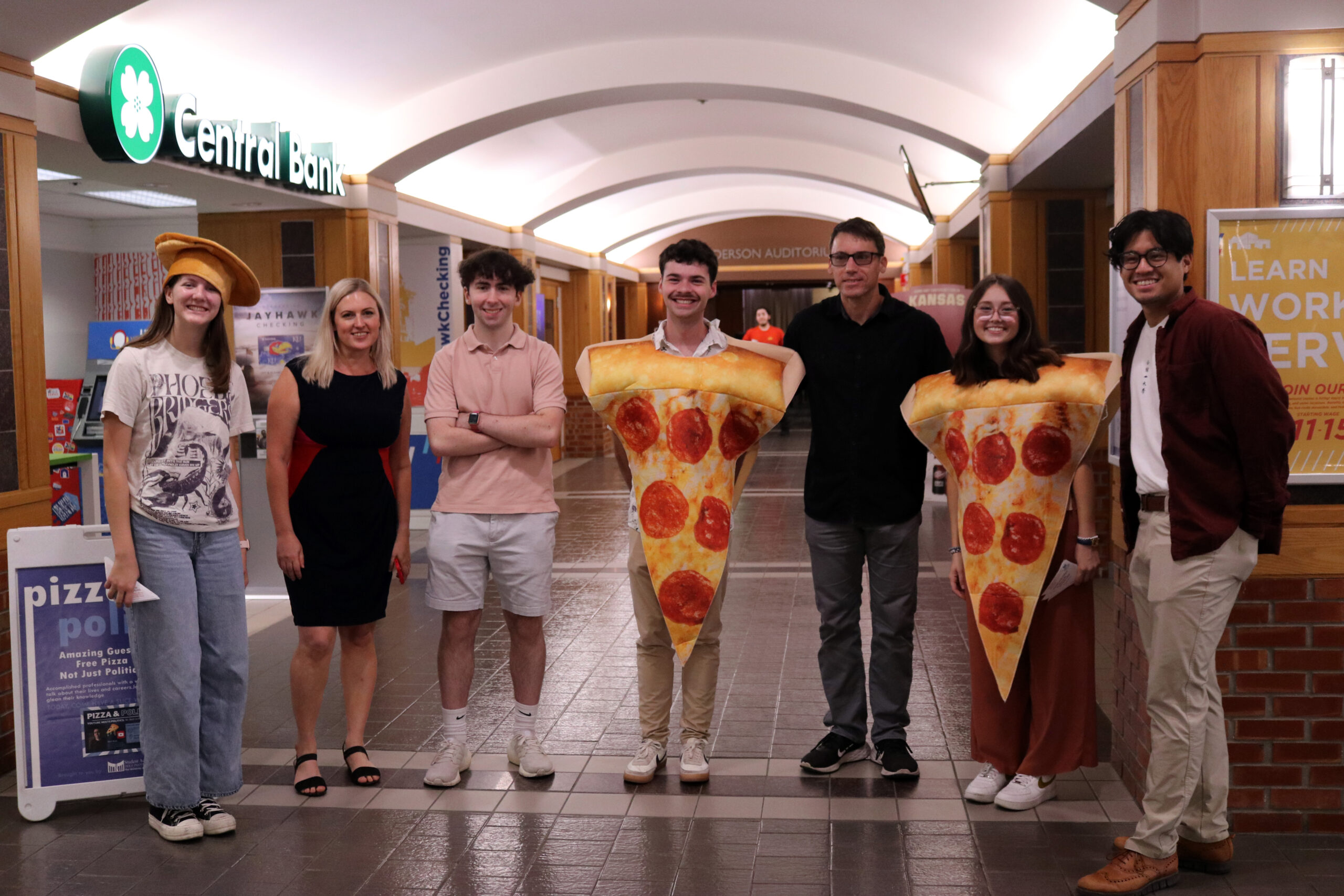 A group of people, some of whom are dressed in a pizza slice costume, pose for a photo in a hallway.