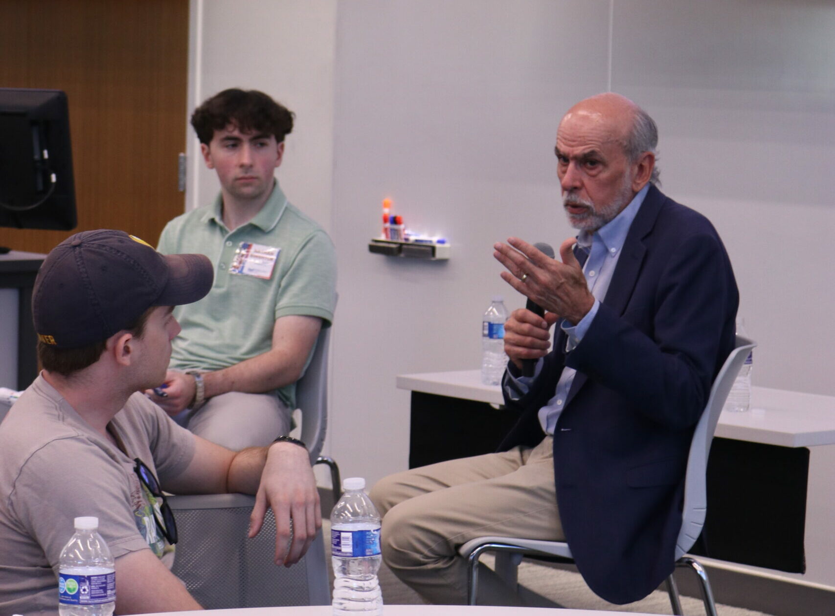 A man in a suit gestures while speaking in a room while people look on.