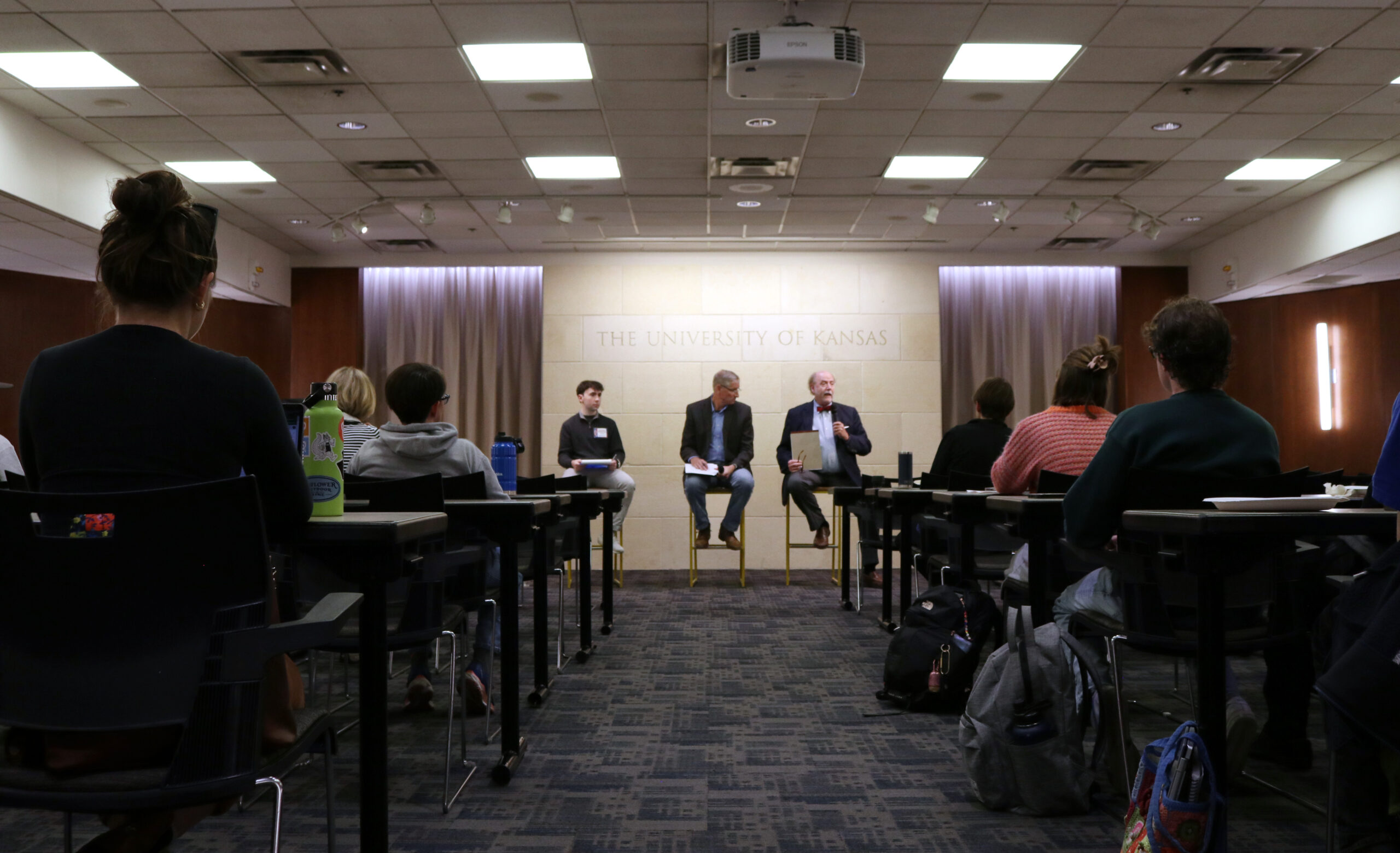 Three men sit in high chairs in the front of a large room while others look on from tables throughout the room.