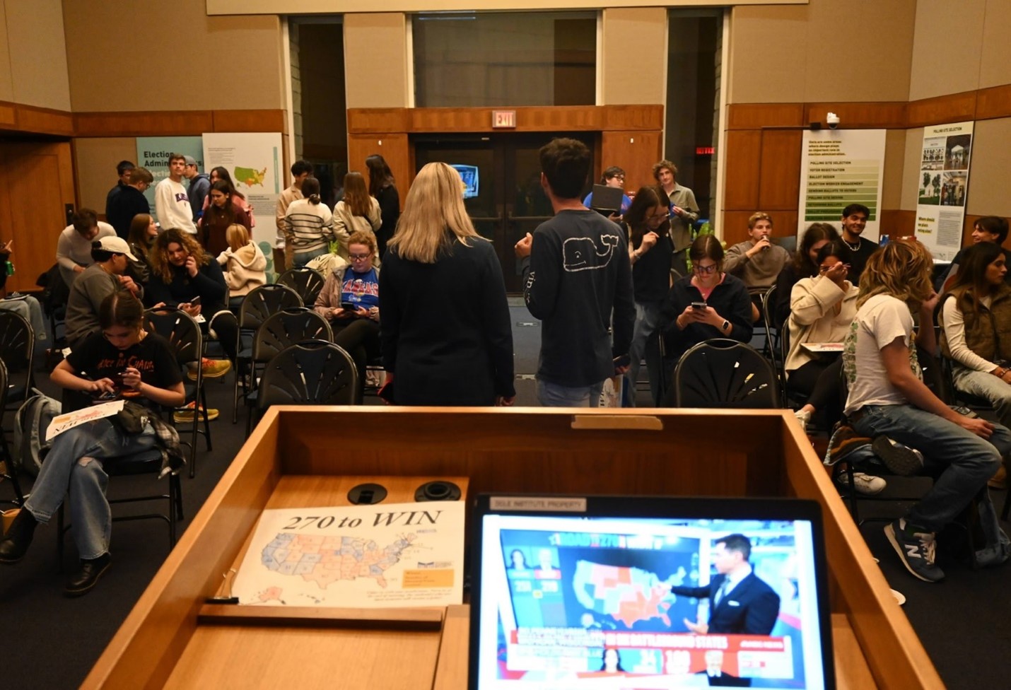 A large group of people sit in a large room while watching election coverage. A podium and laptop are in the foreground.