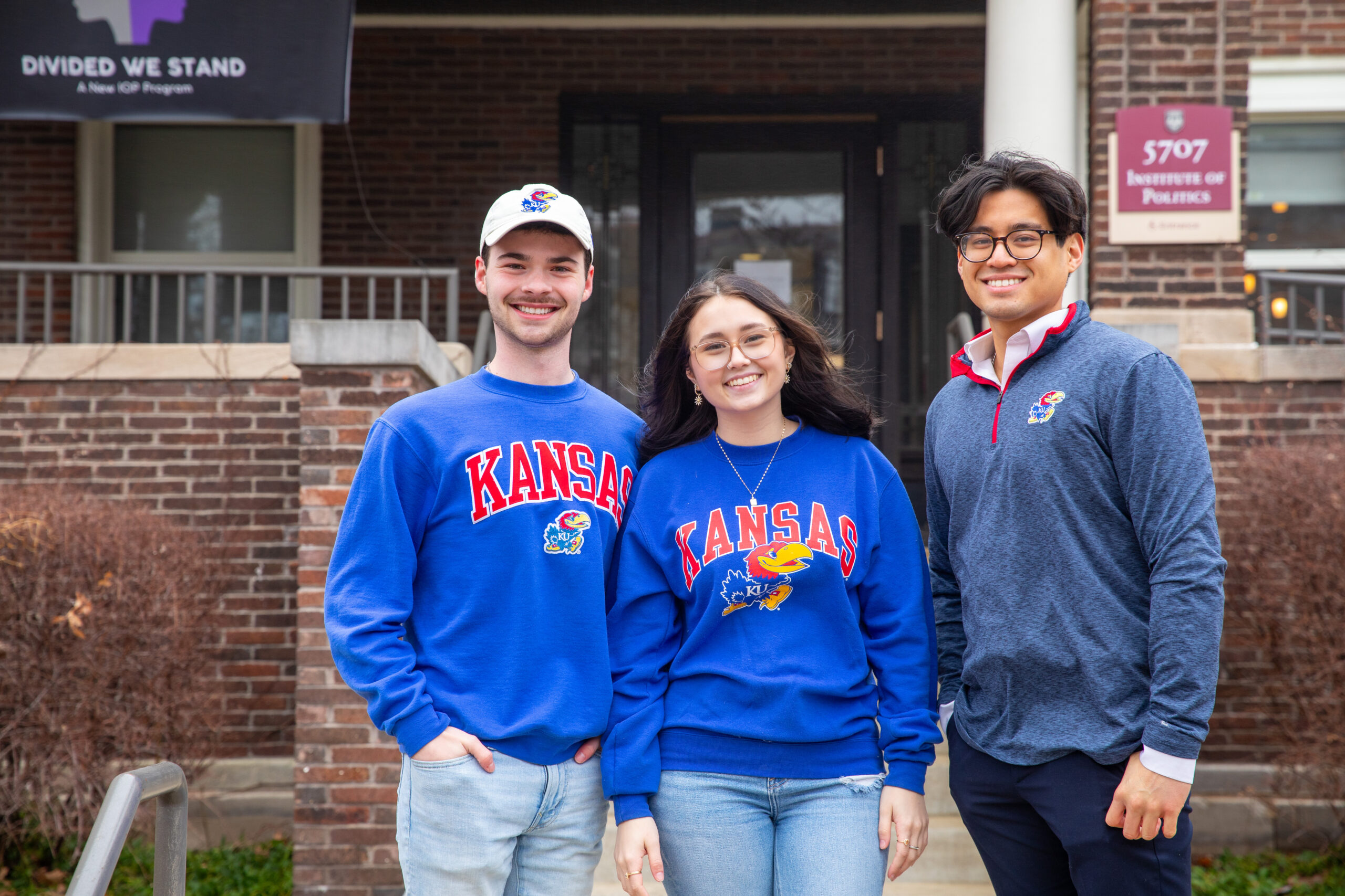Two men and a woman in University of Kansas sweaters stand in front of the front porch of a brick-and-mortar building.