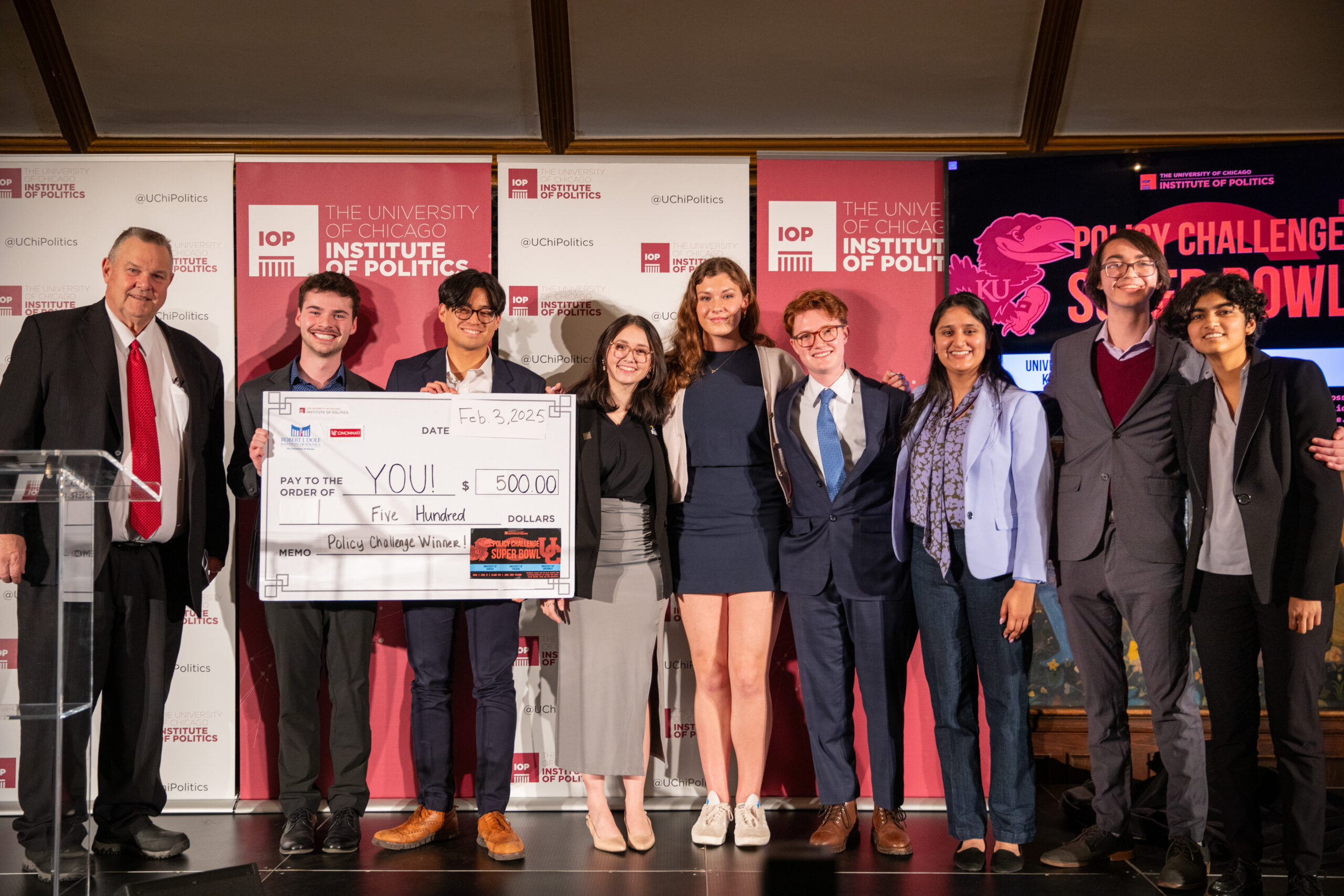 Men and women in business attire stand on a stage for a photograph. Two are holding a giant white check. A red backdrop is behind them.