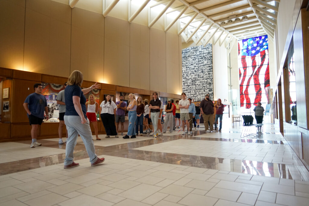 A group of people stands in a spacious, well-lit hallway with high ceilings, large windows, and display cases along the walls. One person gestures toward the group, possibly leading a tour or presentation. A large stained glass window depicting an American flag is visible at the far end of the hallway.